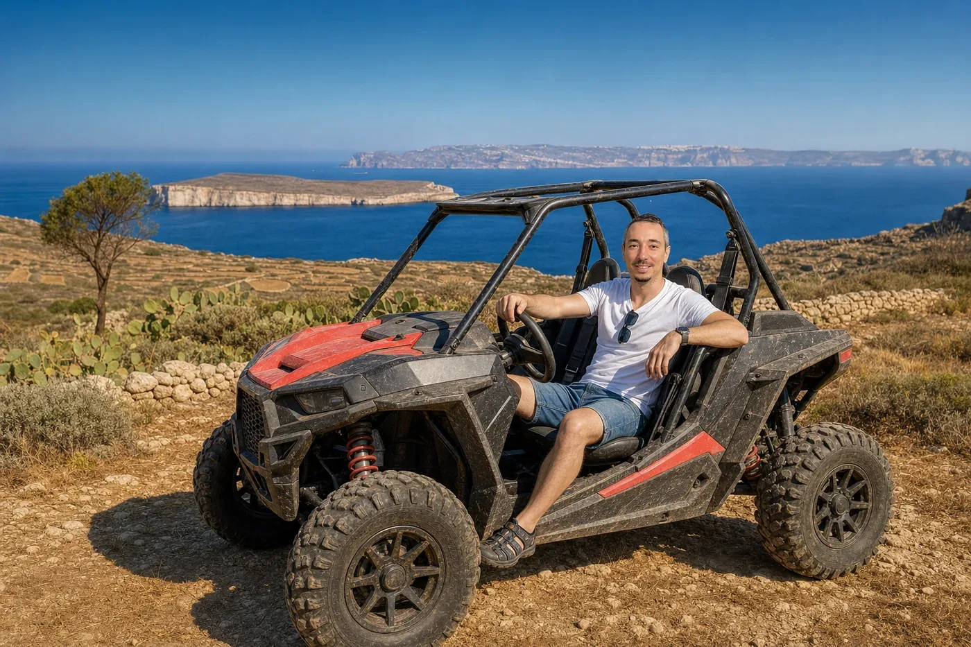 Janez sitting relaxed in a parked off-road buggy on a headland in Gozo, Malta, looking at the camera, with the island of Comino and the coast of Malta across the water behind him